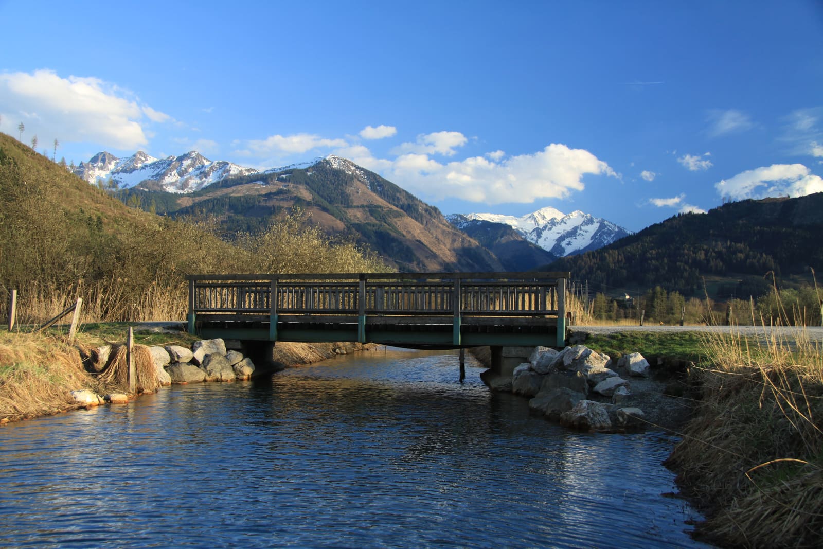 Het vlakke oeverpad bij Zell am See, met een houten brug over het water en besneeuwde toppen erachter.